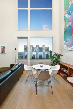 Ein modernes Wohnzimmer mit großem Fenster, einer Couch, einem Esstisch mit Stühlen und einem Bücherregal.