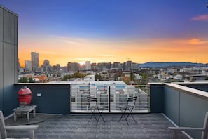 Eine Dachterrasse mit Blick auf die Skyline der Stadt bei Sonnenuntergang, mit Grill, zwei Stühlen und einem Tisch.