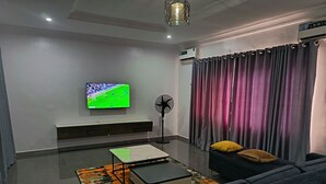 A modern living room with a flat-screen TV, a standing fan, and a sofa. - Kbazzapartments (Ibadan)