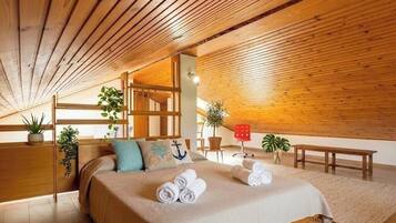 A cozy room with a wooden ceiling, a bed with rolled towels, a bench, and potted plants.