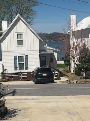 Une maison blanche à deux étages avec un garage, un VUS noir garé devant, et un lac en arrière-plan.