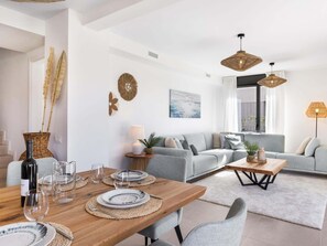 A modern living room with a grey sofa, a wooden dining table set with plates and glasses, and a large window with curtains. - Villa Oronella with Saltwater Pool, Premium Comfort & Elegant Design (Puigderros (Llucmajor))