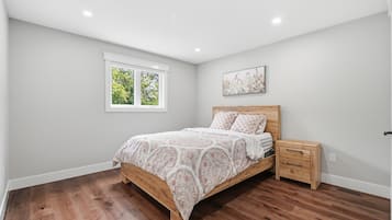 Un dormitorio con cama de madera, mesita de noche, ventana con vista a árboles y un cuadro en la pared.