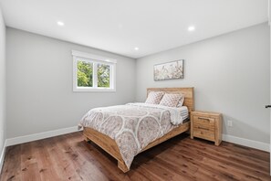 A bedroom with a wooden bed, a nightstand, a window with a view of trees, and a painting on the wall.