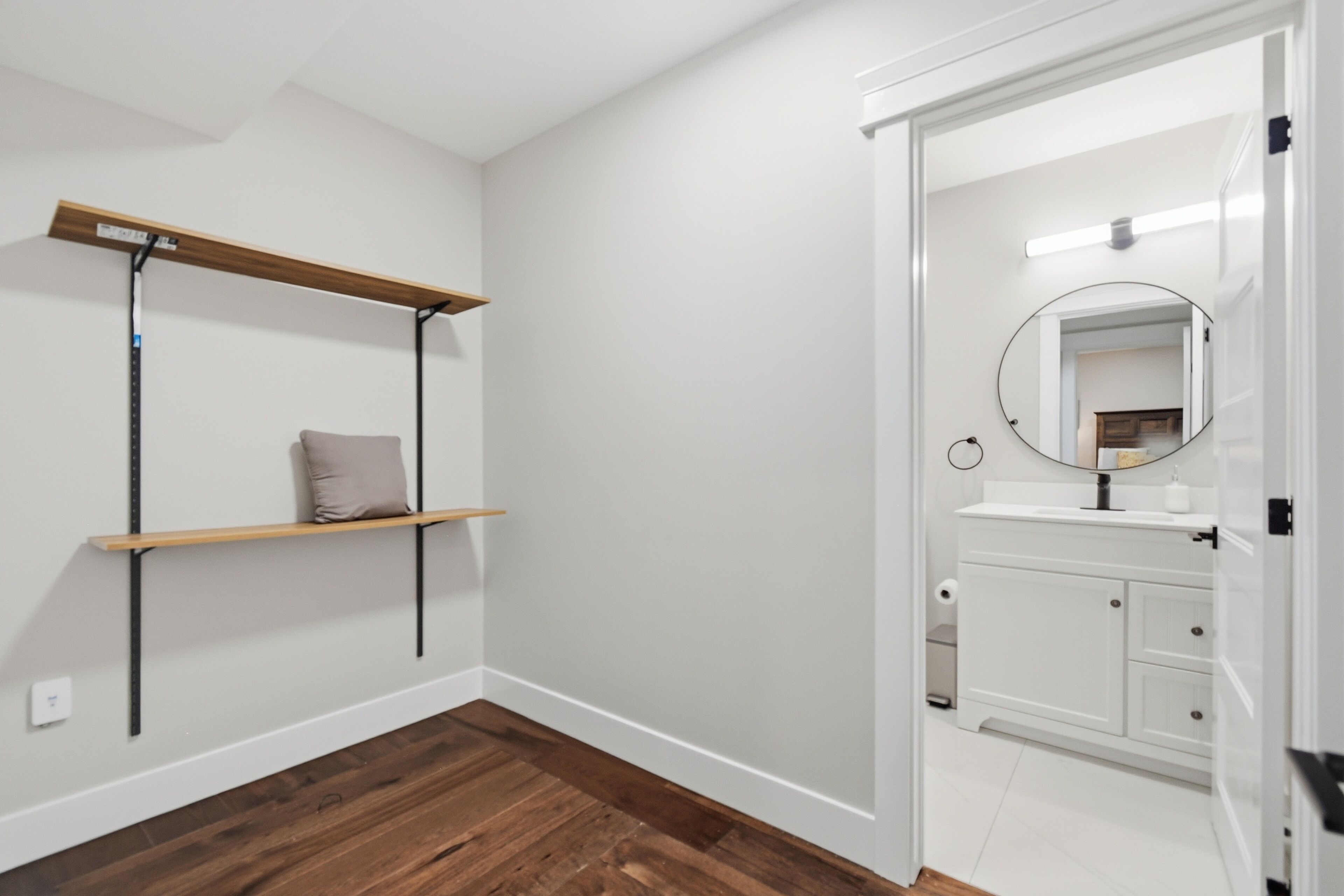 A room with a wooden floor, white walls, a white door leading to a bathroom, and a shelf with a cushion.