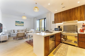 A modern kitchen with wooden cabinets, a built-in oven, and a dining area with a sofa and table. - Chic 2-Bed Apartment with Balcony and Waterfront Views in Port de La Mer (Dubai)