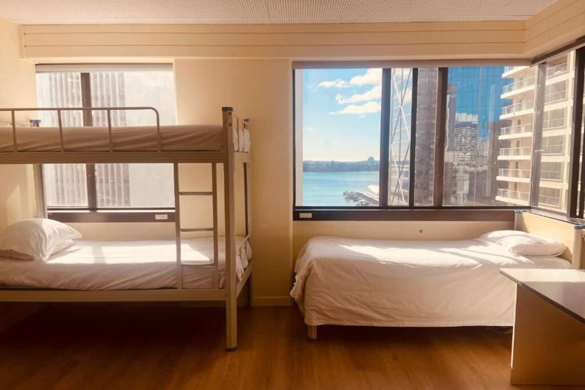 Une chambre avec des lits superposés, un bureau et une grande fenêtre donnant sur un plan d’eau et des bâtiments.