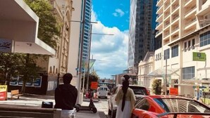 A city street with tall buildings, a red sports car, and a person in a white dress walking. - Garden Hostel (Auckland)