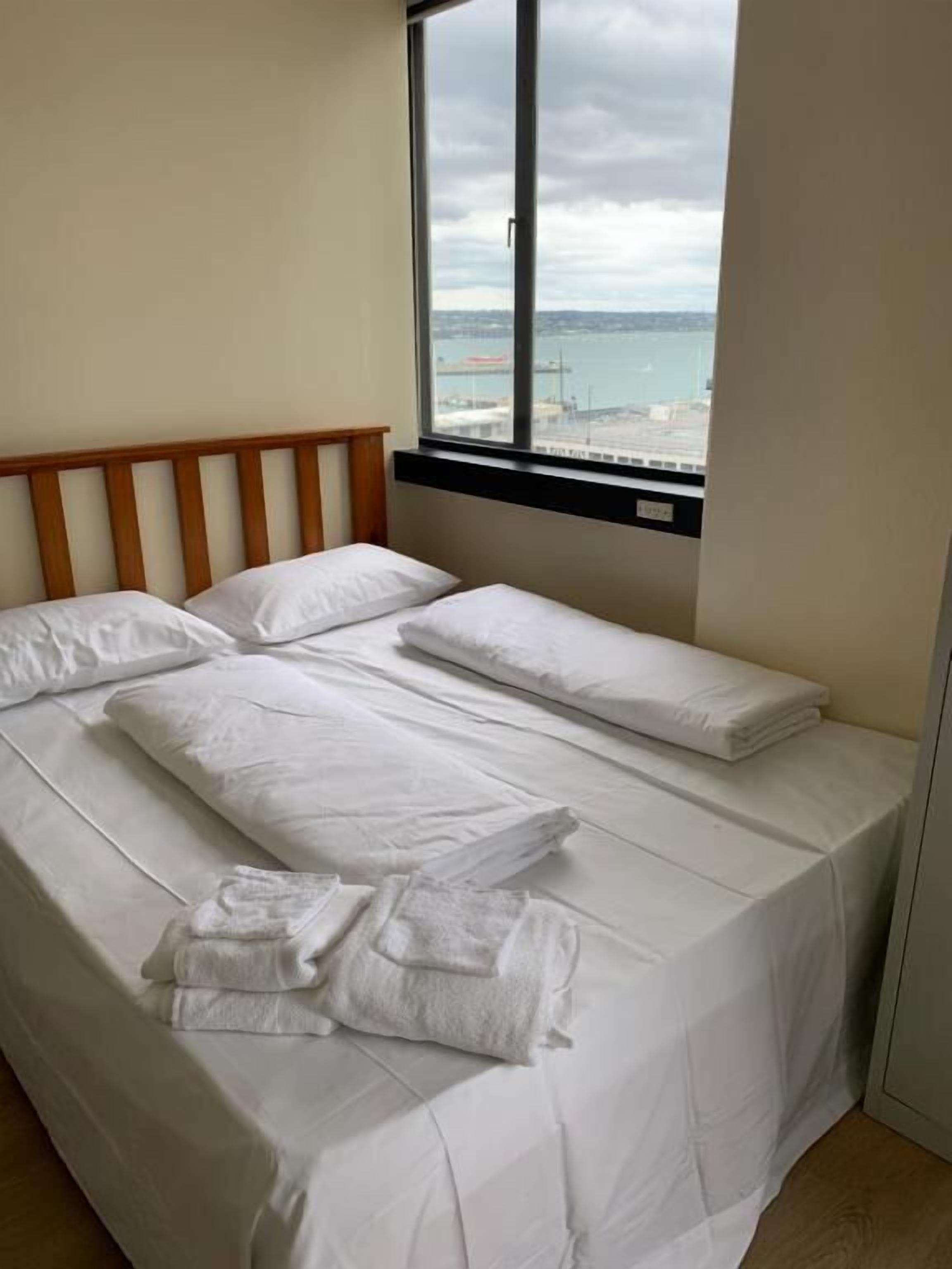 Una cama con sábanas blancas, cabecera de madera y ventana con vista al mar y un puente.