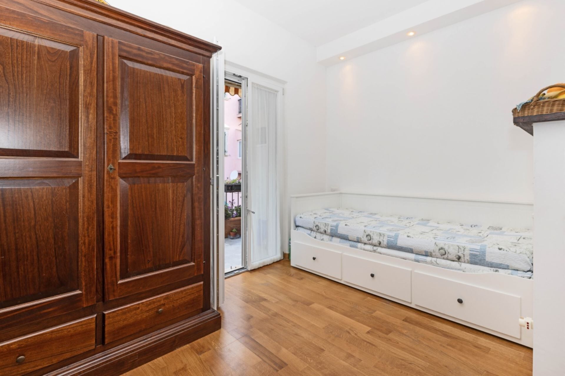 A room with a wooden wardrobe, a white bed with patterned bedding, and a white wall.