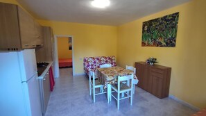A compact kitchen with a refrigerator, stove, and cabinets. There is a dining table with chairs and a bed in the adjacent room. - VILLA PINETA MARE - PIGNA 1 - VILLA PINETA MARE (Capoliveri)