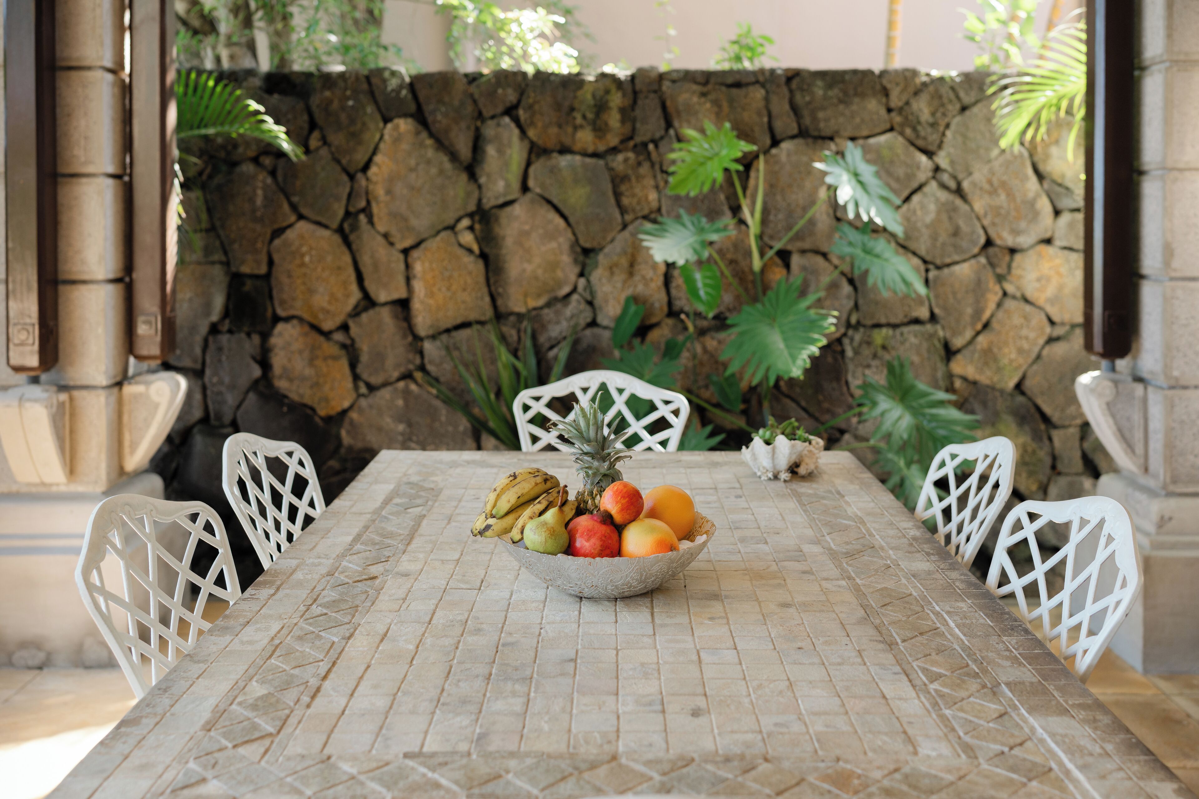 Un mur de pierres, une table sur laquelle repose un panier tressé rempli de fruits, et des chaises blanches.