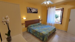 Een slaapkamer met een bed, een plafondventilator, een raam met gordijnen, een aan de muur hangend schilderij en een kleine potplant.