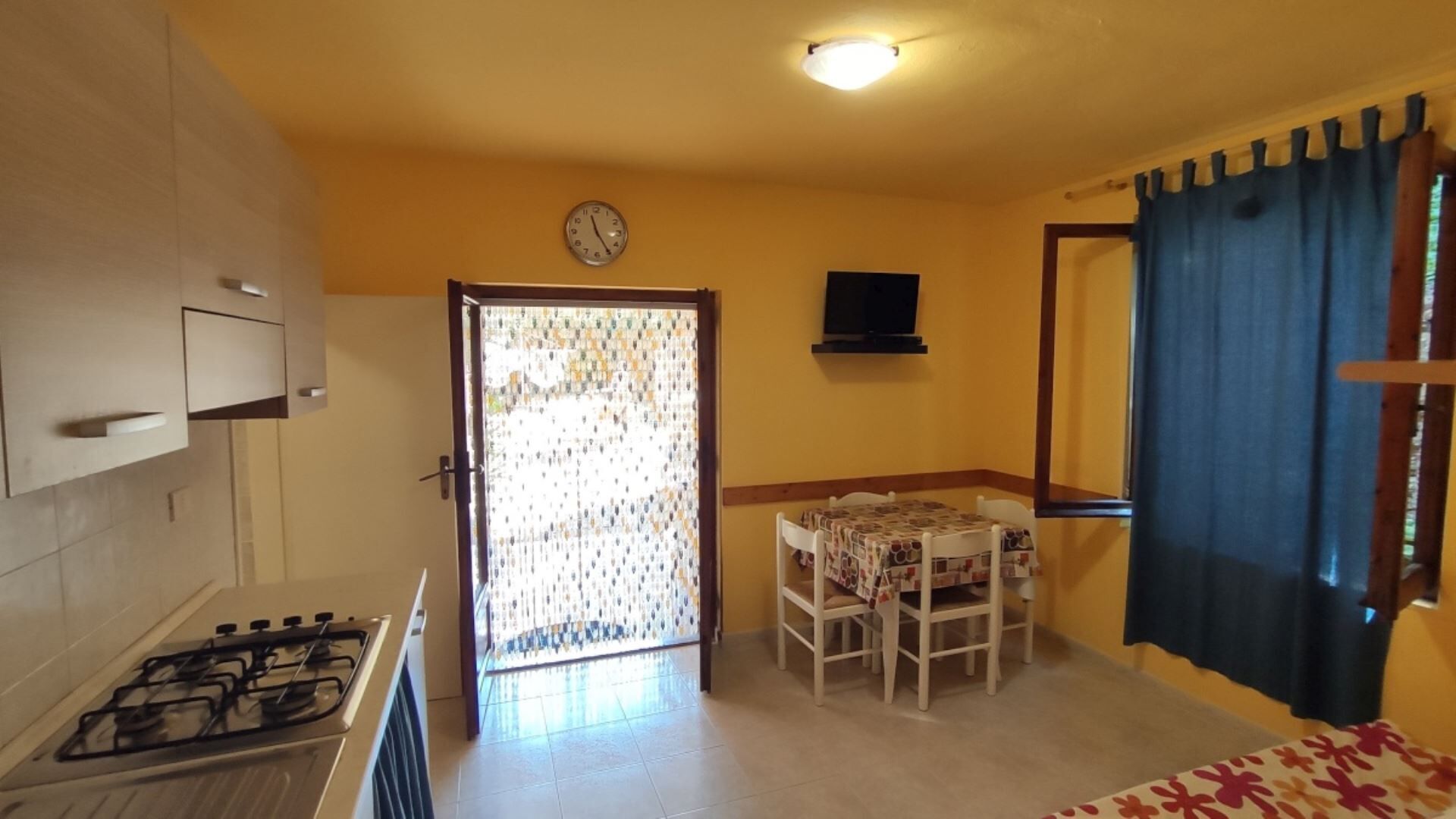 A compact kitchen with a gas stove, a dining area with a table and chairs, a clock on the wall, and a door with patterned curtains.