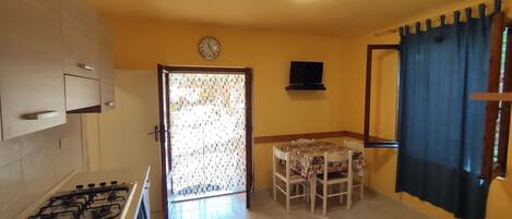 A compact kitchen with a gas stove, a dining area with a table and chairs, a clock on the wall, and a door with patterned curtains.