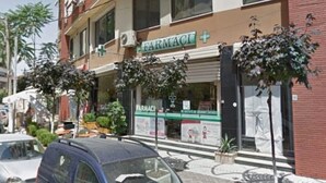 Un edificio con letrero de farmacia, un coche azul estacionado adelante y un pequeño árbol en una jardinera.