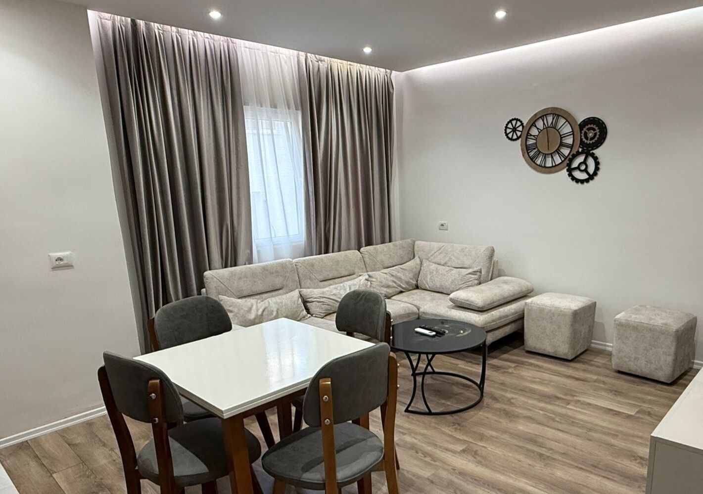 Een moderne woonkamer met een hoekbank, een eettafel met stoelen, een klein rond tafeltje en een decoratieve wandklok.