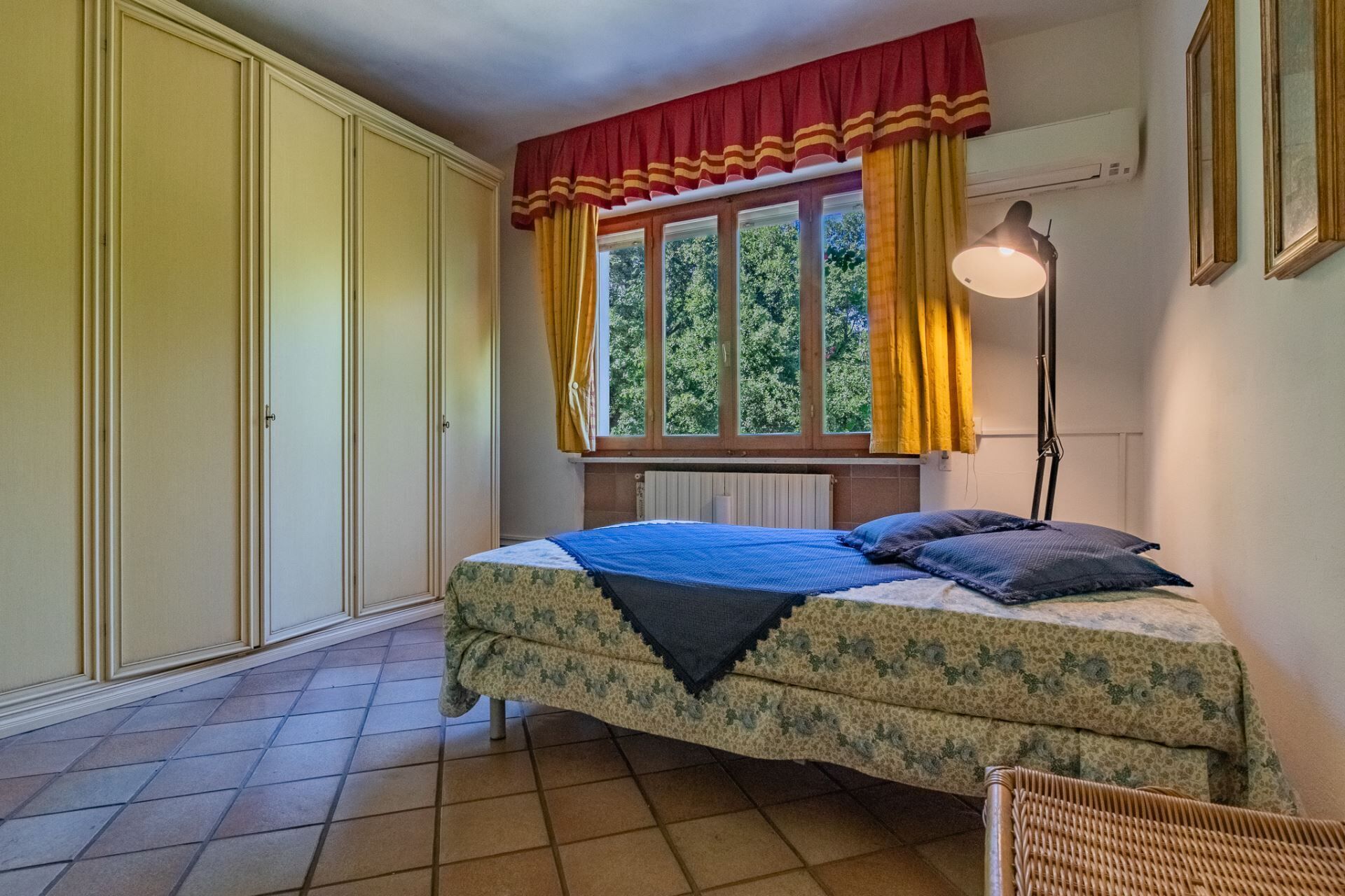 Un dormitorio con cama, ventana con cortinas, radiador y lámpara.