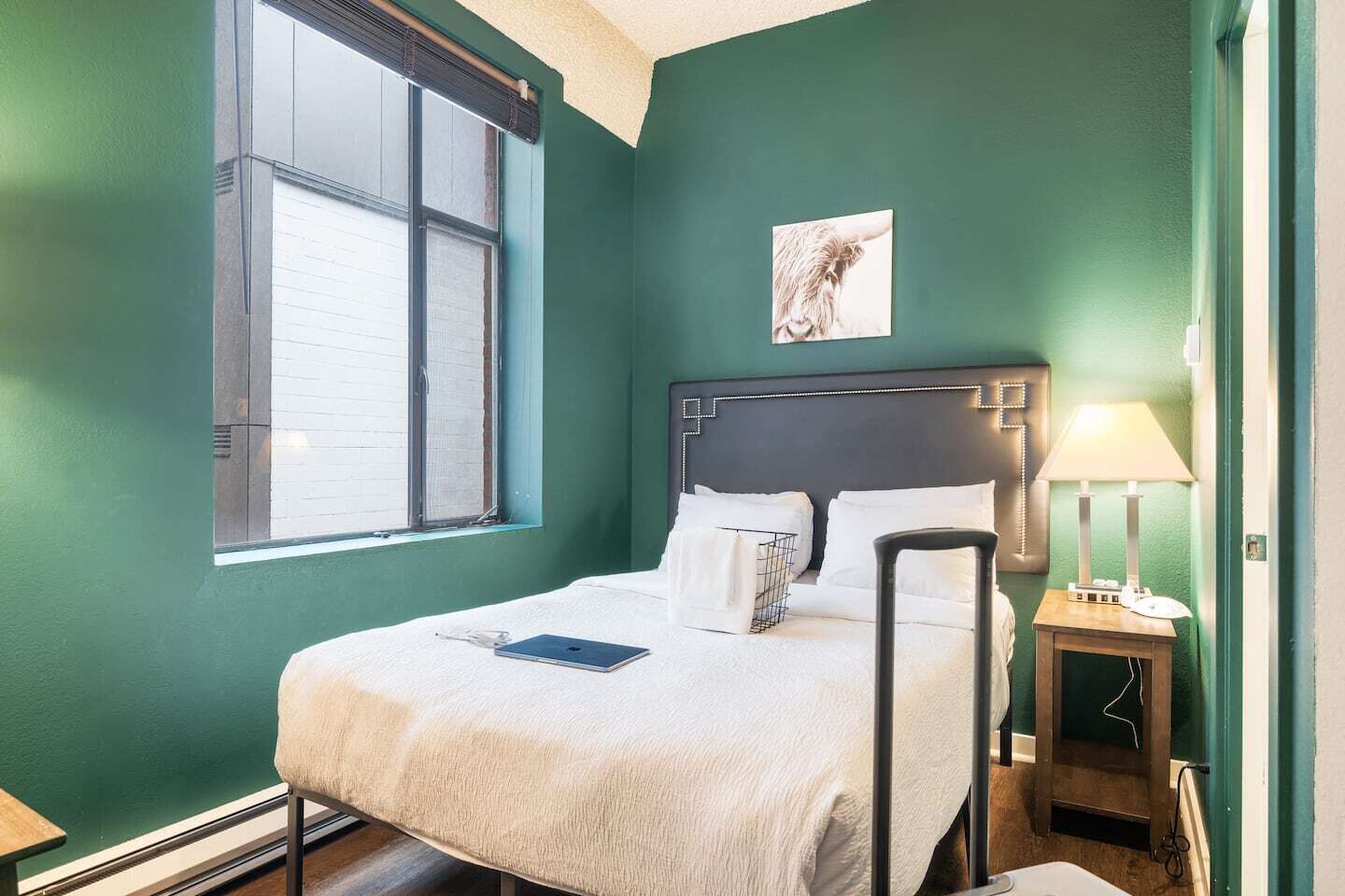 A neatly made bed with white linens, a bedside table with a lamp, a window with blinds, and a framed picture on the wall.