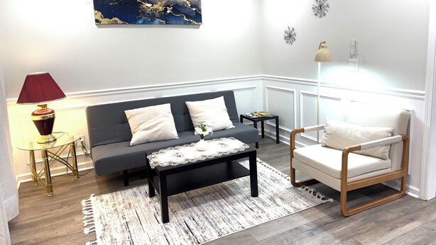 Una sala de estar con un sofá gris oscuro, un sillón blanco, una mesa de centro, una lámpara roja y un elemento decorativo en la pared.