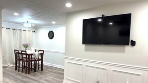 Un comedor con mesa y sillas de madera, un reloj en la pared y un televisor de pantalla plana montado encima.