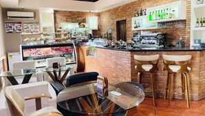 Un café avec un mur en briques, un coin bar avec des tabourets, une vitrine remplie de pâtisseries et un ventilateur au plafond.