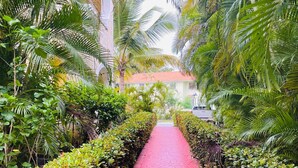 Un chemin pavé de briques rouges, bordé d’une végétation luxuriante et de palmiers, mène à un bâtiment surmonté d’un toit recouvert de tuiles rouges.