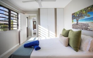Ein Schlafzimmer mit einem Bett, Kissen, einem Fenster und einem Gemälde, das eine Strandansicht zeigt.