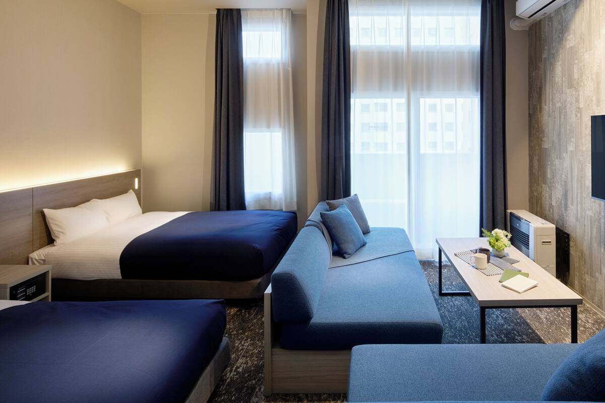 A hotel room with two beds, a sofa, a desk with a flat-screen TV, and a window with curtains.