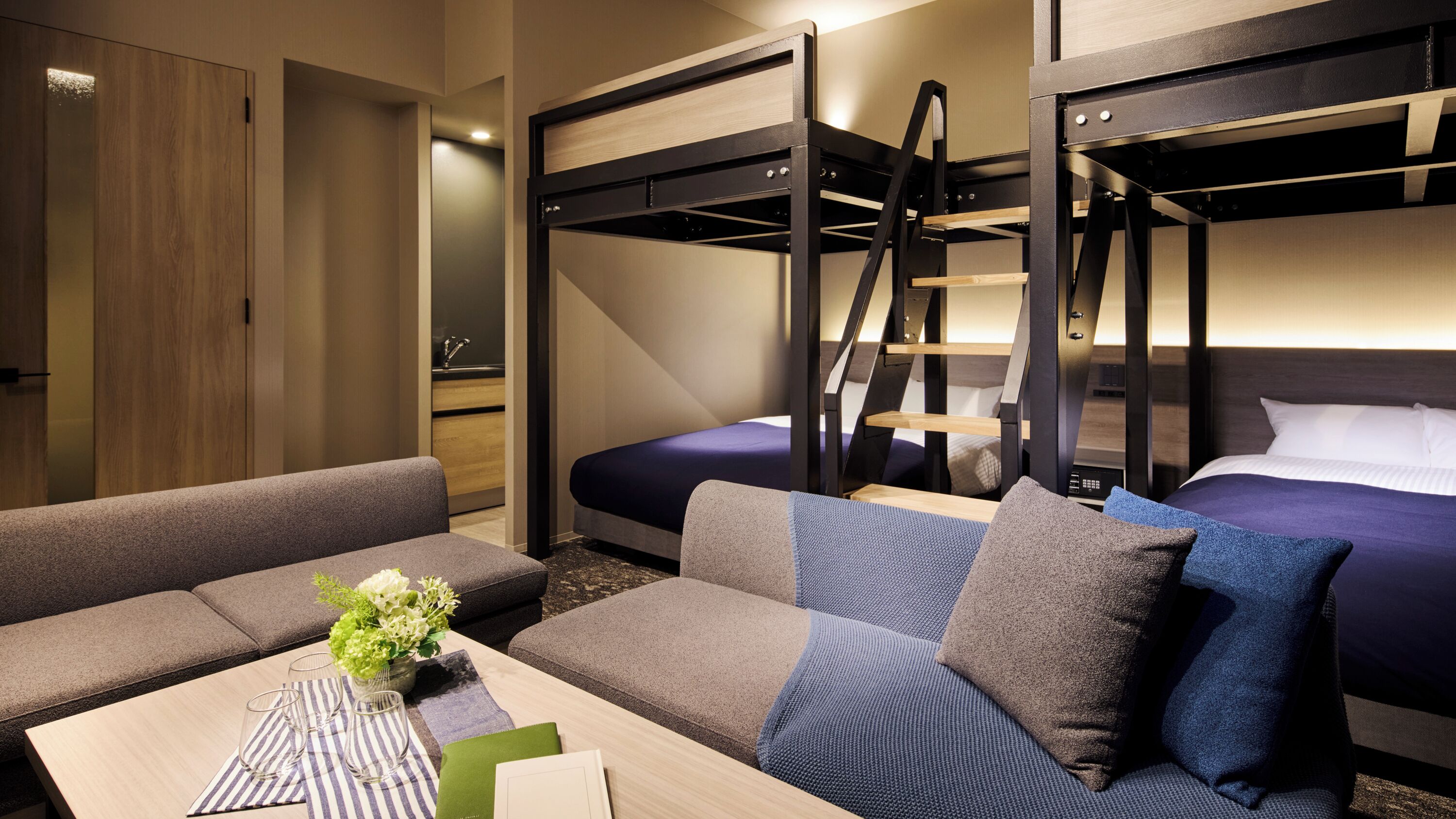 A hotel room with a bunk bed, a sofa, a small table with a vase of flowers, and a bedside table with a book and a glass.