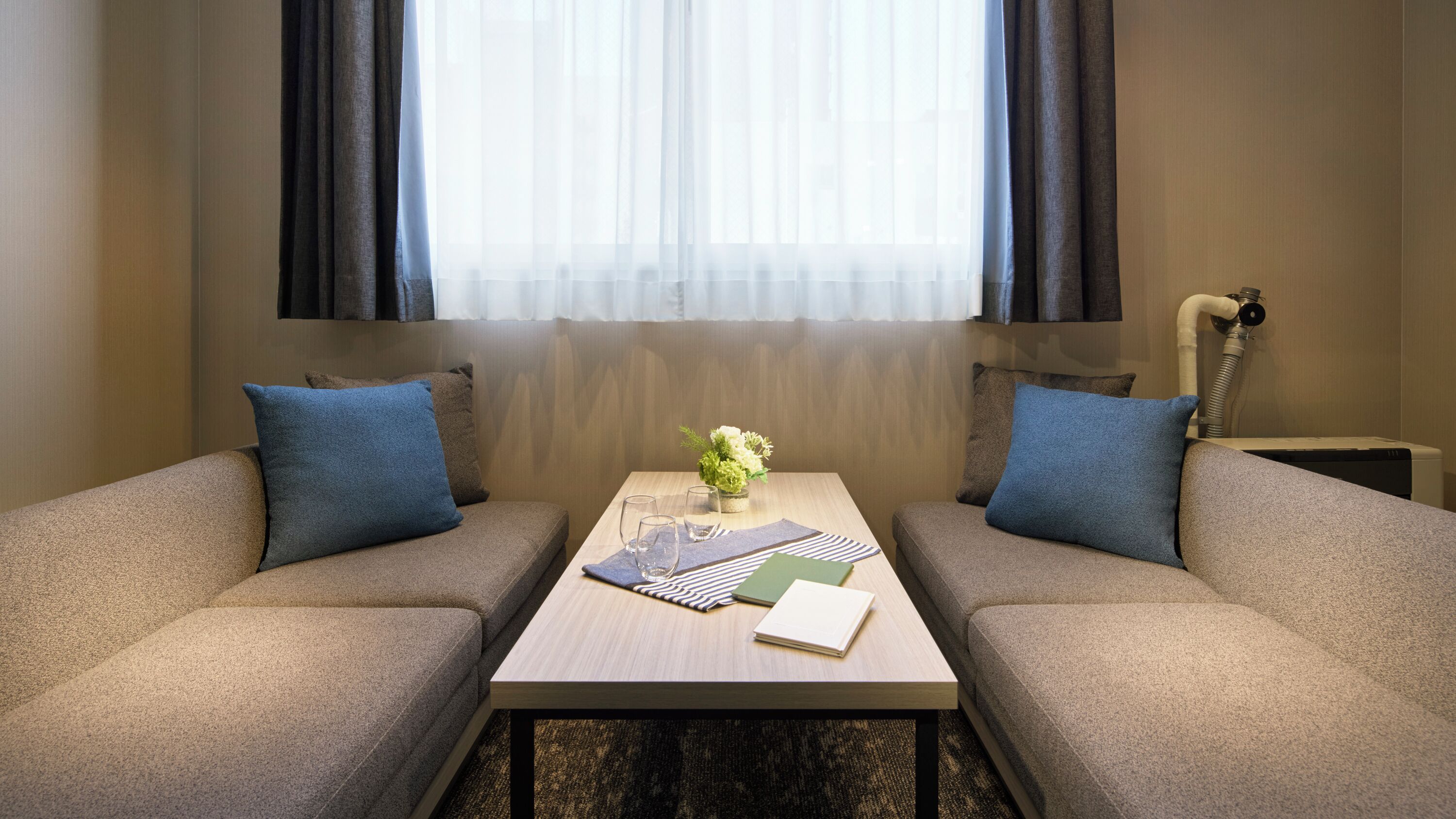 Hotellihuone, jossa on kaksi sohvaa, pieni pöytä maljakko täynnä kukkia ja ikkuna, jossa on verhot.