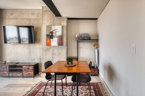 Una sala de estar moderna con una mesa de comedor de madera, sillas negras, un televisor montado en una pared de concreto y una cama tipo loft con una manta.