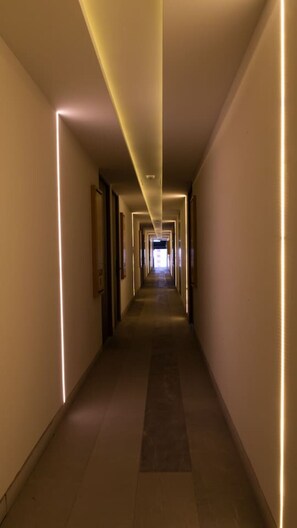 A long, narrow hotel corridor with a central light fixture and framed pictures on the walls.