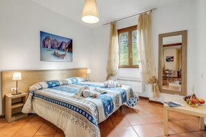 A bedroom with a bed, nightstands, a window with curtains, a painting, and a table with a bottle and fruit. - Residence La Perla - Apt 17 (Monte Isola)