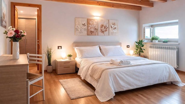A bedroom with a wooden ceiling, a bed with white bedding, a nightstand with a lamp, a chair, a small table with a vase of flowers, and a window with a view. - Nuvolau Appartement Dolomites (Perarolo di Cadore)