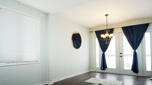 Ein Zimmer mit dunklem Holzboden, weißen Wänden, einem runden Spiegel, einer Chandelüster und einem großen Fenster mit Vorhängen.