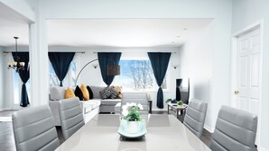 Ein geräumiges Wohnzimmer mit einem großen Esstisch, grau gepolsterten Sesseln, einem grauen Sofa mit gemusterten Kissen und Blick ins Freie.