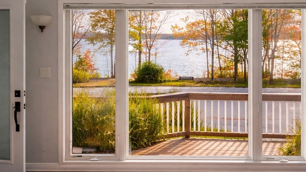 Ein Zimmer mit einer großen Schiebeglasdoor, die auf eine Terrasse mit Blick auf einen See und Herbstlaub führt.