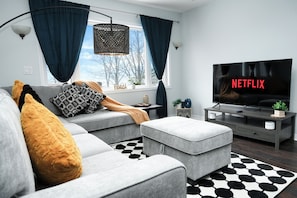 A modern living room with a grey sofa, ottoman, and a flat-screen TV displaying the Netflix logo. - Luxury Lakeview Stay with Jacuzzi | Downtown Barrie (Barrie)