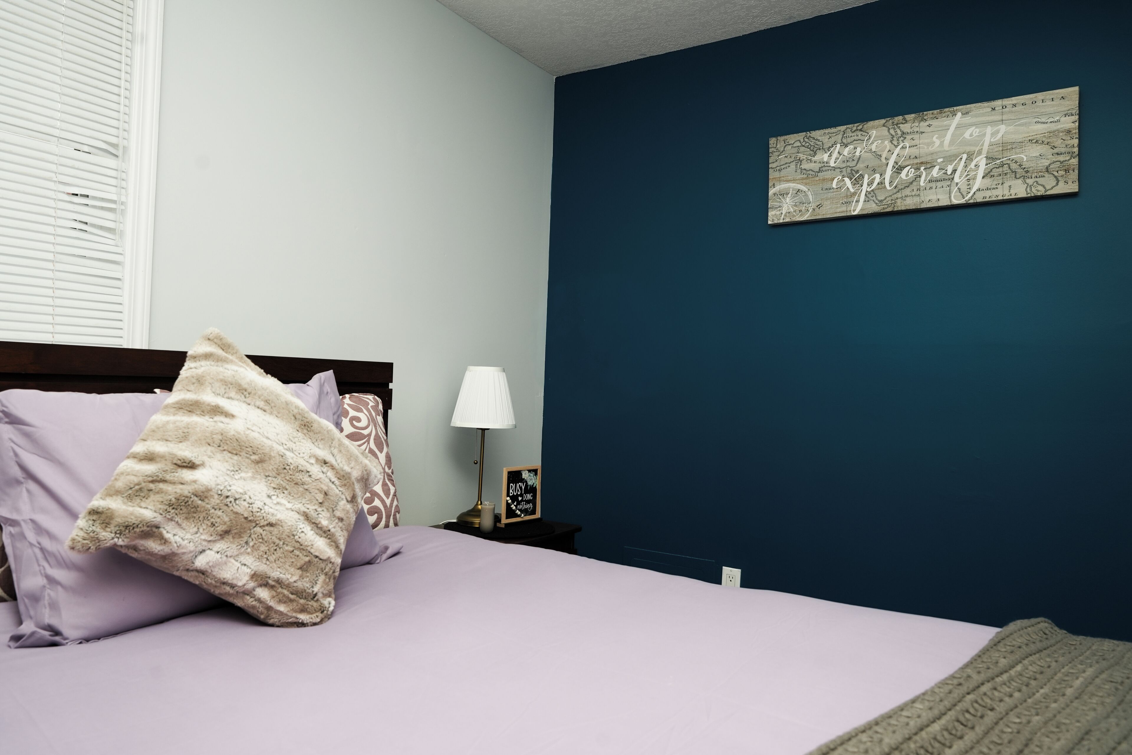 Un dormitorio con cama, almohadas, una mesita de noche con lámpara y una pared con un mapa y una cita motivacional.
