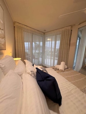 Une chambre d’hôtel avec un grand lit, un coin salon avec un fauteuil et une vue dégagée à travers des rideaux légers.