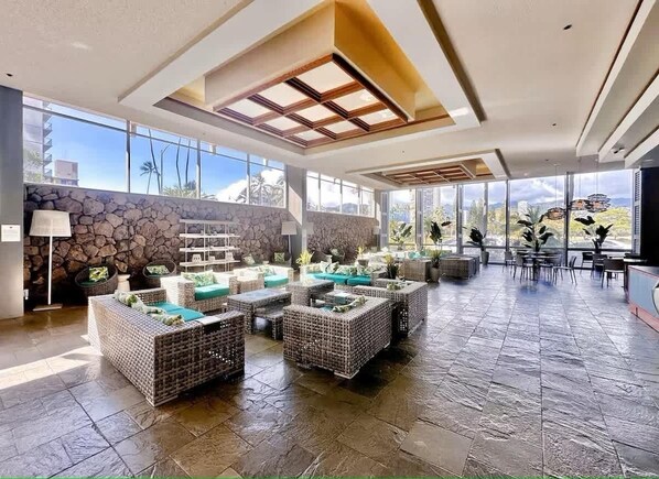 A spacious lobby with wicker furniture, stone walls, and large windows offering views of palm trees and buildings.