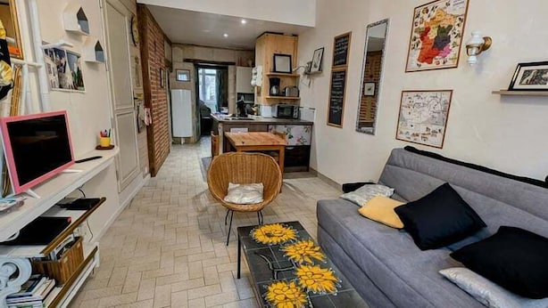 Een moderne woonkamer met een grijze bank, een bijzettafeltje met gele bloemen, een rieten fauteuil en een keuken op de achtergrond.