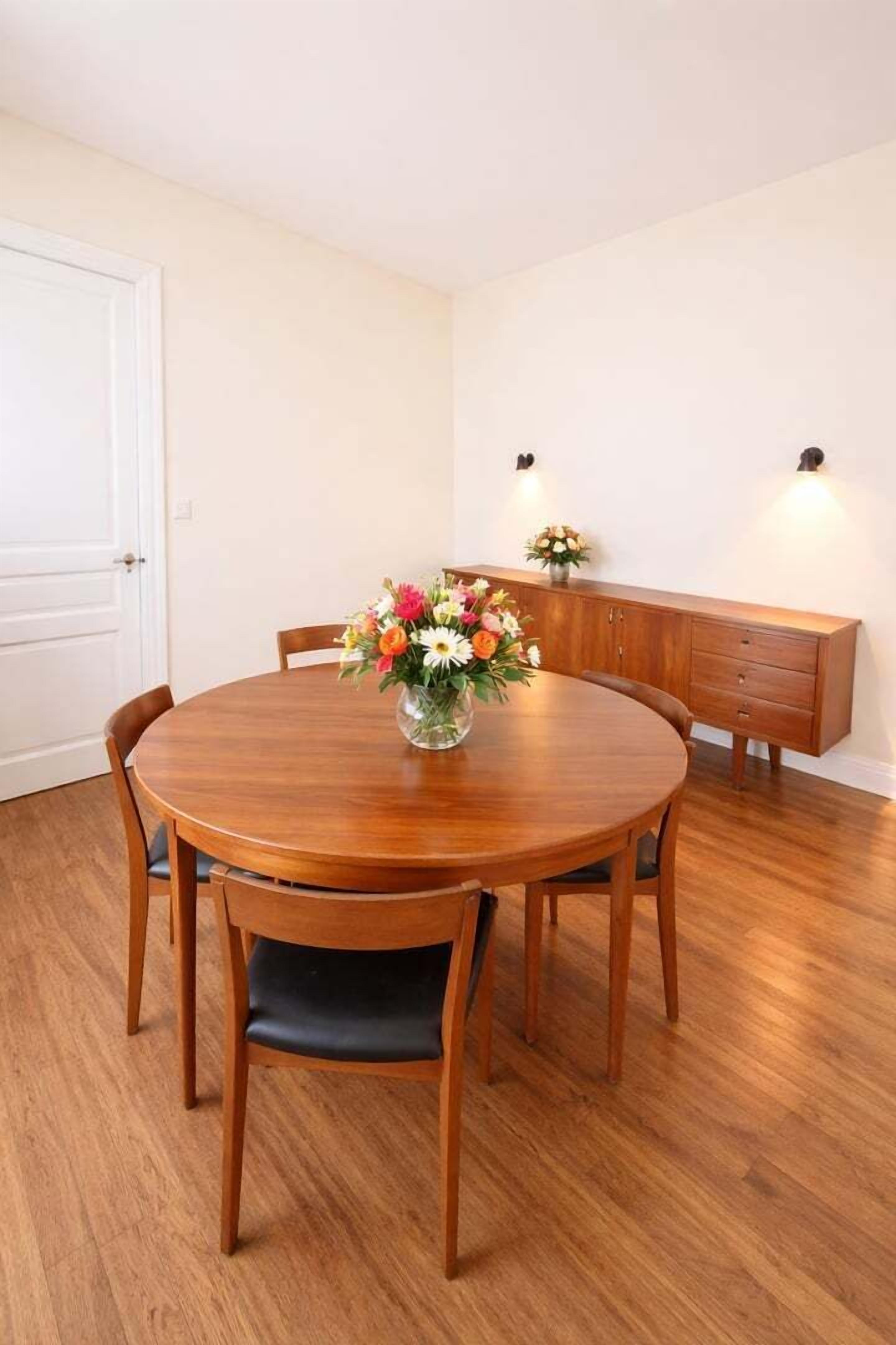Un comedor con una mesa de madera, sillas, un jarrón con flores y un aparador de madera.