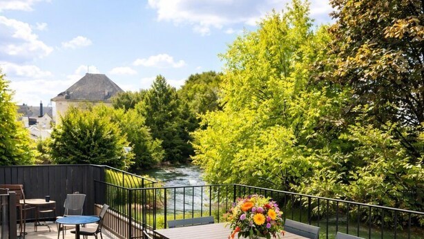 A wooden deck with a table and chairs, a flower vase, and a view of a river and trees.