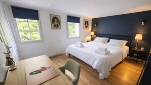 A hotel room with a bed, two bedside lamps, a table with a chair, and two windows.