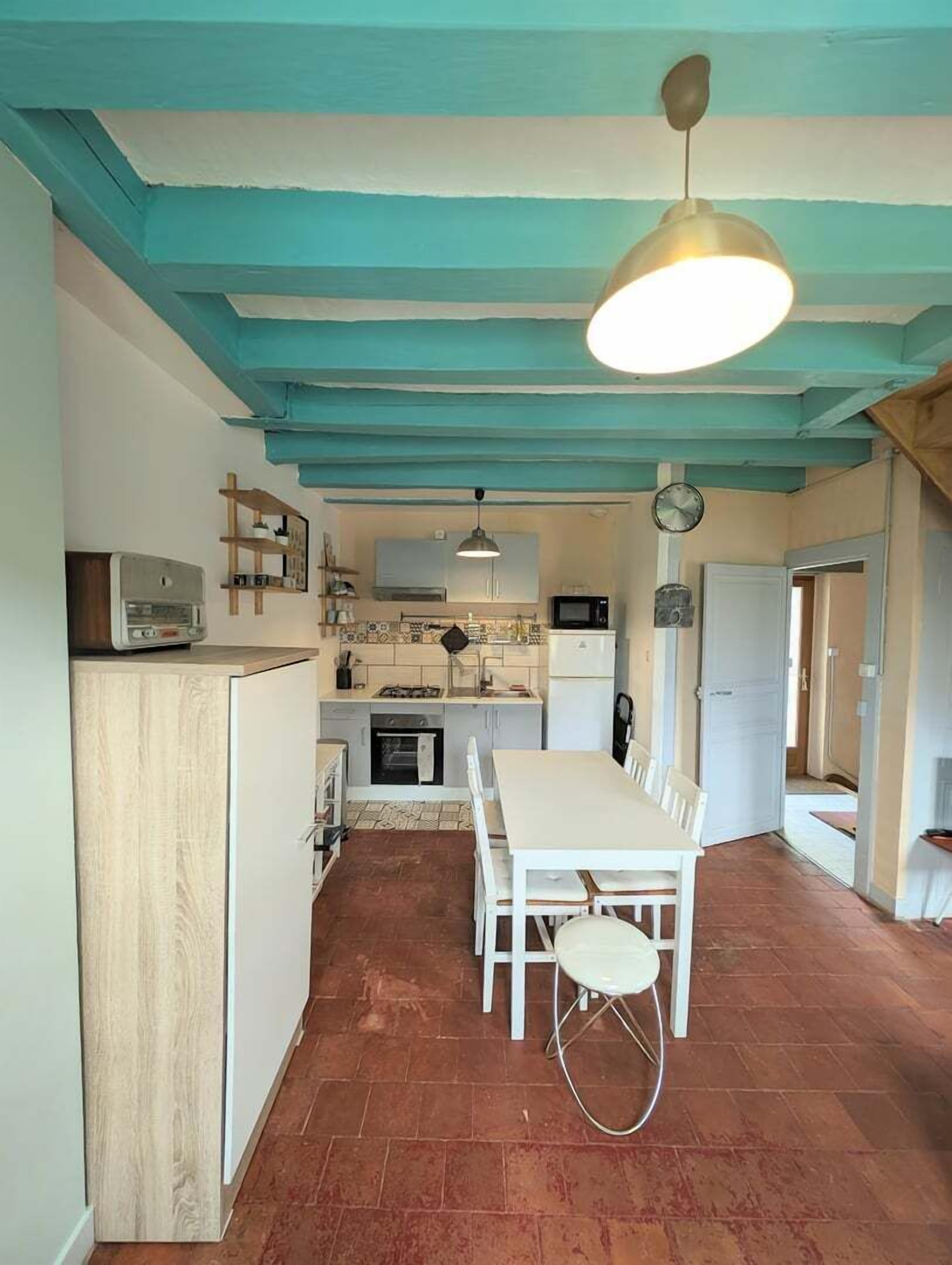 A compact kitchen with a dining area, featuring a white table and chairs, a hanging light fixture, and a clock on the wall.