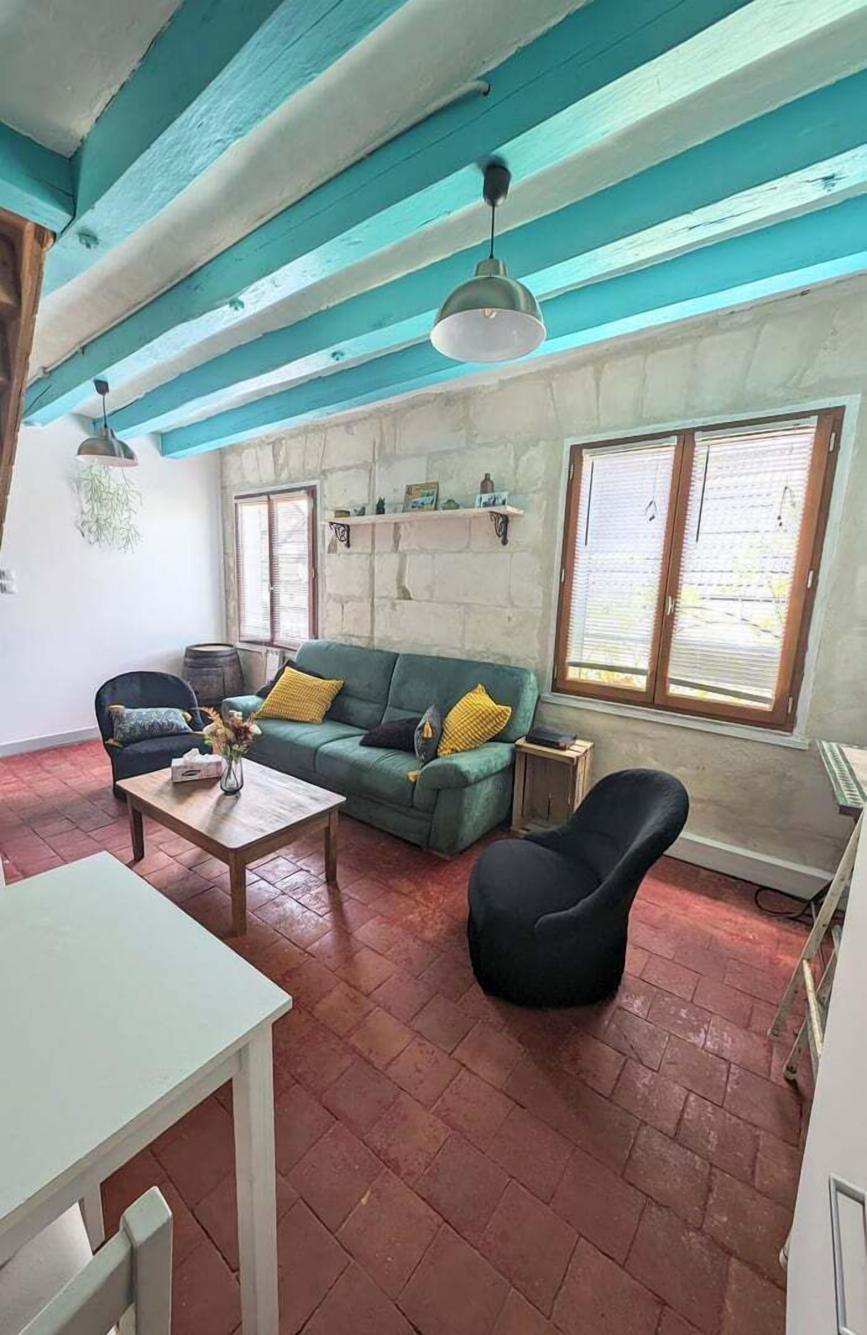 A living room with a green sofa, a black armchair, a wooden coffee table, and a white table. There are two hanging lamps, a window with blinds, and a stone wall.
