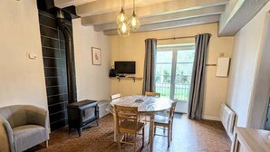 A room with a hexagonal tile floor, a round dining table with chairs, a sofa, a TV mounted on the wall, and a window with curtains. - The source - Le Grand-Pressigny (Le Grand-Pressigny)