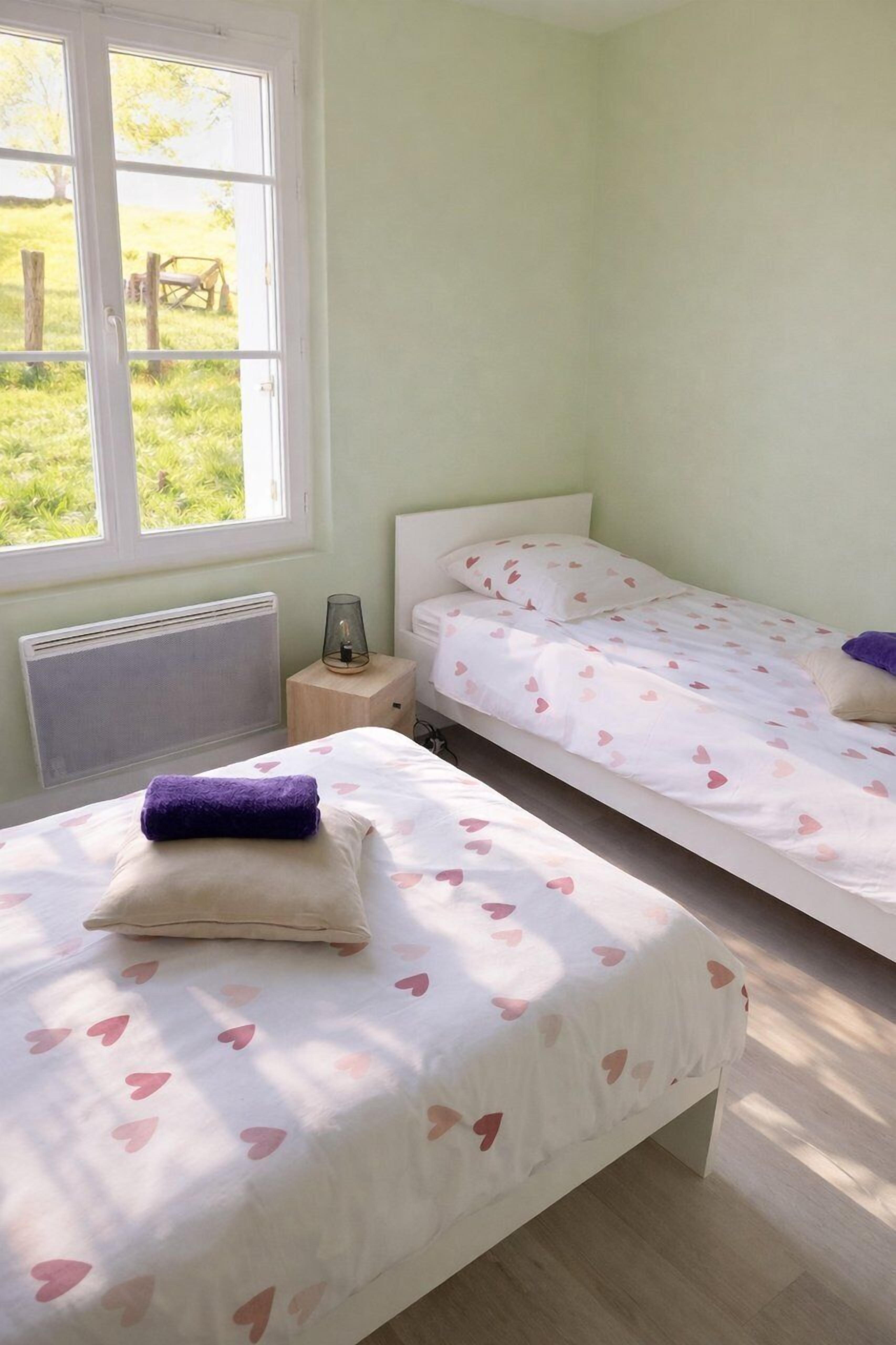 A bedroom with two beds, a radiator, a bedside table with a lamp, and a window with a view of the outdoors.